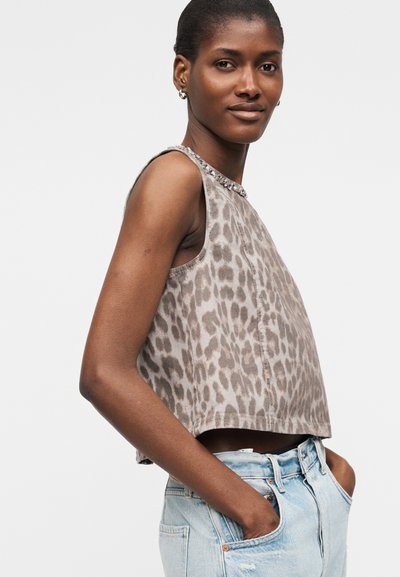 Γυναίκα που φοράει αμάνικη γκρι crop top με λεοπάρ σχέδιο και διακοσμητικό λαιμόκοψη, ανοιχτό μπλε ψηλόμεσα τζιν, με τα χέρια στις τσέπες, σε λευκό φόντο.