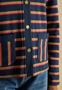 Námornícky kardigán s horizontálnymi pruhmi v zlatej a ružovej farbe. Má zlaté gombíky a dve predné vrecká pre pohodlné ukladanie.