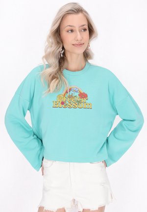 Világoskék pulóver, oversized ujjakra tervezve, virágos grafikával, amelyen a "Feelin' Blossom" felirat található. Megfelelően viselt fehér, kopott rövidnadrággal.