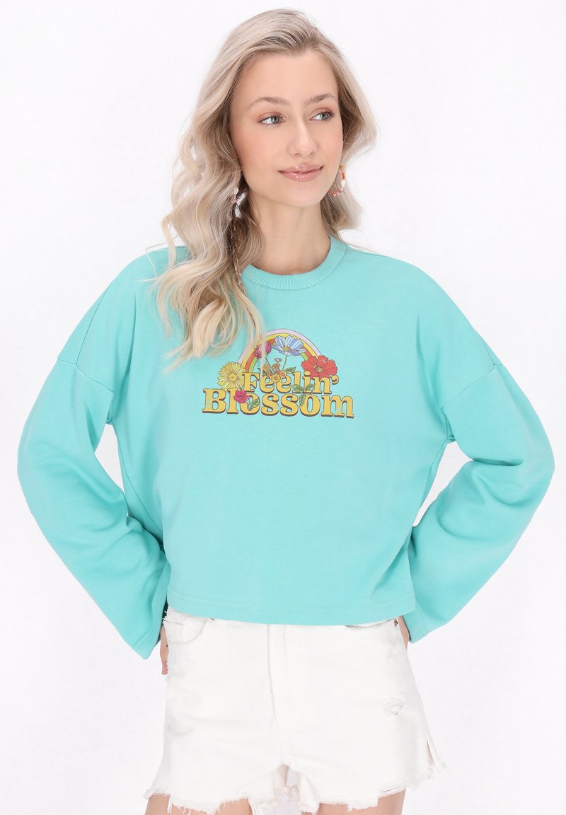 Világoskék pulóver, oversized ujjakra tervezve, virágos grafikával, amelyen a "Feelin' Blossom" felirat található. Megfelelően viselt fehér, kopott rövidnadrággal.