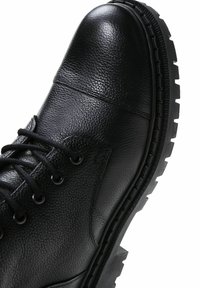 Botín de cuero negro con una superficie texturizada, puntera redonda, diseño de cordones y una suela gruesa de goma con tacos prominentes.