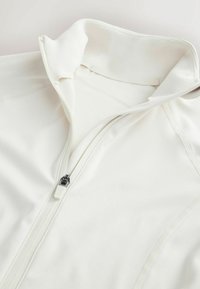 Veste en tissu blanc doux avec une fermeture éclair partiellement ouverte et un col relevé, montrant des coutures détaillées sur un matériau lisse.