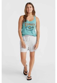 Türkises Tanktop mit grafischem Text, kombiniert mit hellgrauen Shorts mit Kordelzug. Model trägt schwarze Flip-Flops und steht vor einem neutralen Hintergrund.