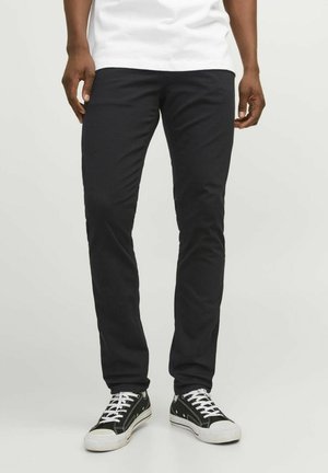 Personne portant un pantalon noir slim et des baskets en toile noire avec des lacets blancs, debout devant un fond clair uni.