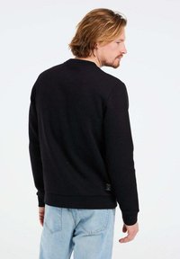 Felpa nera con scollo a girocollo, polsini e orlo a costine, materiale in cotone, design semplice e piccolo dettaglio con etichetta vicino alla cucitura inferiore.