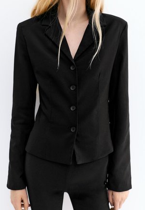 Vrouw met een nauwsluitend zwart blazer met vier knopen en bijpassende zwarte broek, van nek tot heupen getoond tegen een witte achtergrond.