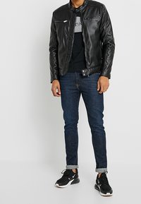 Chaqueta de cuero negra con bolsillos con cremallera, llevada sobre una camiseta negra y gris. Jeans ajustados azules y zapatillas deportivas negras con detalles en blanco.