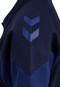 Marinblå pikétröja i texturerat tyg, med en chevron-mönster i en ljusare blå nyans på axeln. Krage med dragkedja.