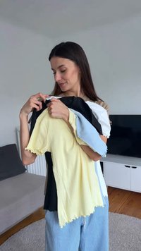 Νεαρή γυναίκα κρατά και κοιτάζει μια στοίβα από διπλωμένα T-shirts σε διάφορα χρώματα μέσα σε ένα σαλόνι.