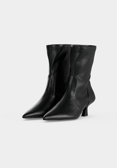 Paire de bottes en cuir noires mi-mollet avec bouts pointus et petits talons blocs sur un fond clair uni.