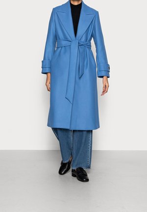 Femme portant un manteau bleu ceinturé longueur genou sur un jean large et des mocassins en cuir noir, debout devant un fond gris uni.