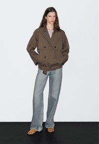 Brown double-breasted jacket with black buttons, worn over a gray top. Paired with light blue flared jeans and brown strappy sandals.