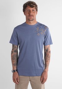 Homme avec des tatouages sur les deux bras portant un t-shirt bleu avec un design floral jaune sur l'épaule gauche et un pantalon beige.