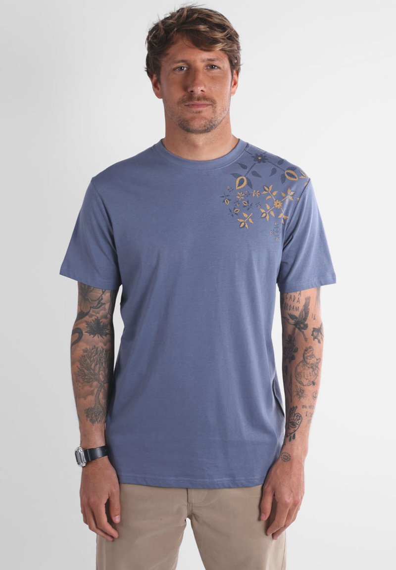 Homme avec des tatouages sur les deux bras portant un t-shirt bleu avec un design floral jaune sur l'épaule gauche et un pantalon beige.