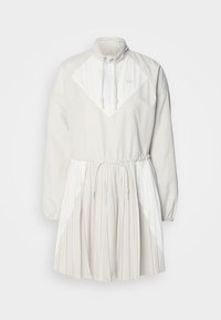 Vit klänning med veckad kjol och dragsko i midjan. Den har en hög krage och dragkedja fram, med kontrasterande triangulära designelement.