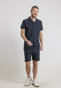 Chemise courte à manches bleu marine avec une poche poitrine, associée à un short assorti. Tissu mélangé coton, fermeture à boutons, et baskets blanches.