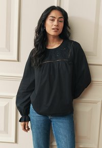 Melnā garajaka blūze ar izšūtu kakla daļu, brīva piegriezuma, ar savilkuma detaļu zem krūtīm, kombinēta ar zilām džinsu biksēm.