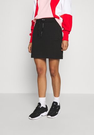 Person iført en sort knælængde nederdel med snore, hvide sokker, sorte sneakers og en rød- og hvidlangærmet top.