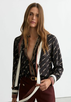 Frau mit langem braunem Haar trägt eine gemusterte schwarze Bluse mit weißem und braunem Besatz, kombiniert mit hoch taillierten weinroten Hosen und einem schwarzen Gürtel.