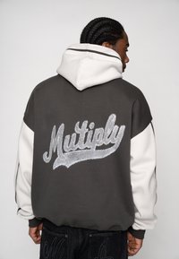 Hanorac gri cu glugă și mâneci albe. Prezintă broderia "Multiply" în argintiu pe spate. Material moale, croială lejeră, manșete cu margine nervurată.