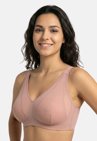 Uśmiechnięta kobieta z falistymi ciemnymi włosami, ubrana w jasnoróżowy bezprzewodowy biustonosz z szerokimi ramiączkami, na tle jednolitego jasnego tła.