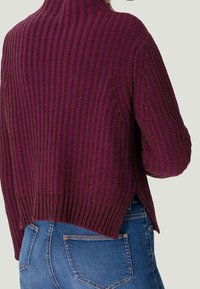 Pull en tricot côtelé bordeaux avec un col montant, des fentes sur les côtés et un tissu texturé. Associé à un jean bleu.