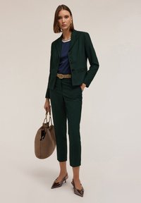 Mörkgrön skräddarsydd kostym med en figursydd kavaj, tillsammans med en marinblå topp, beige bälte, brun handväska och blanka spetsiga klackskor.