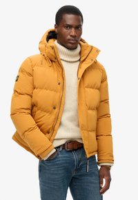 Giacca a vento gialla con cappuccio, texture trapuntata, chiusure con zip e bottoni, abbinata a un dolcevita crema e jeans blu.