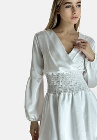 Witte satijnen jurk met een gerimpelde taille, lange opgeblazen mouwen en een diepe V-halslijn, met een vloeiende rok. Gladde textuur.