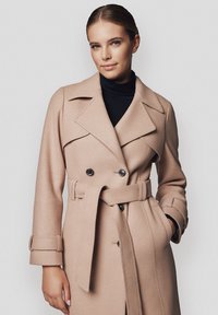 Beżowy płaszcz dwurzędowy wykonany z mieszanki wełny, z paskiem w talii, szerokimi klapami i dekoracyjnymi guzikami; gładka tekstura i dopasowany krój.