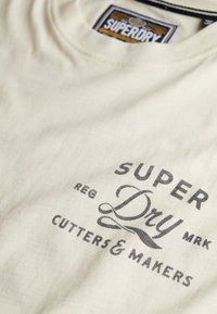 Camiseta de algodón color crema con cuello redondo y texto impreso en negro: "SUPER DRY CUTTERS & MAKERS" en el frente.