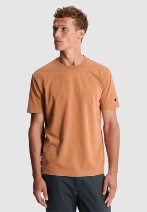 Homme aux cheveux bouclés portant un t-shirt uni orange-marron à manches courtes et un pantalon sombre, regardant vers sa gauche sur un fond clair.
