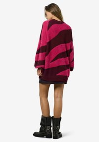 Liels dzijas džemperis rozā un bordo krāsā ar drosmīgām zigzaga svītrām. Tam ir teksturēta auduma un platās piedurknes. Valkāts virs melnām svārkiem.