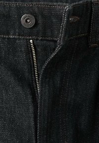 Vaqueros de mezclilla negros con cremallera y cierre de botón, con costuras en contraste y textura suave. Cremallera y cierre metálico visibles.