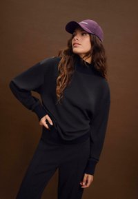 Felpa nera con un collo alto e polsini a coste, abbinata a pantaloni neri. Un berretto viola con la scritta "New York".