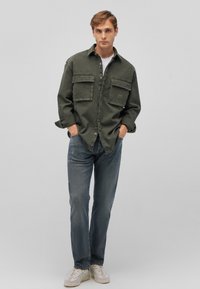 Olivgrünes Denim-Hemd mit zwei vorderen Taschen und geknöpften Bündchen. Über einem weißen T-Shirt getragen und kombiniert mit blauen Jeans und weißen Turnschuhen.