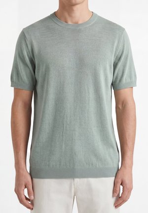 Homme portant un pull en maille vert clair à manches courtes avec col, poignets et ourlet côtelés, associé à un pantalon beige clair.