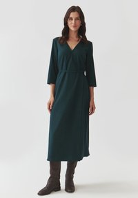 Robe longue verte enroulée en tissu doux, avec un décolleté en V profond, des manches trois-quarts et une taille nouée, associée à des bottes marron montantes jusqu'aux genoux.