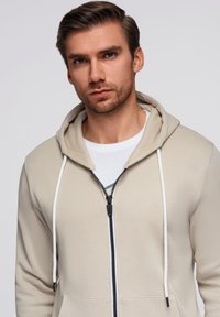 Felpa beige con zip e cappuccio regolabile, dotata di zipper nera e dettagli bianchi. Realizzata in tessuto morbido, progettata con una vestibilità comoda.