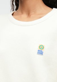 Fehér pulóver bordázott kereknyakkal és színes "House of Colors" logóval a bal oldalon. Puha pamut textúra.