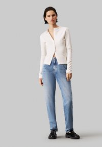 Cárdigan blanco de canalé con cuello redondo y cierre de botones, combinado con jeans rectos de color azul claro y zapatos negros.