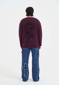 Bordowy sweter dzianinowy z czarnym wzorem smoka na plecach. W połączeniu z niebieskimi dżinsowymi spodniami w paski z aplikacjami i grafikami.