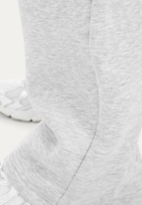 Lichtgrijze joggingbroekspijp met zichtbare naad, gecombineerd met een witte sportschoen op een lichte achtergrond.