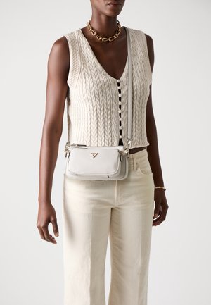 Femme portant un haut sans manches crème en maille torsadée, pantalon crème, collier chaîne en or, bracelet et petit sac bandoulière blanc.