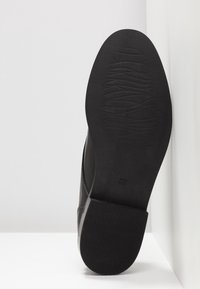 Semelle d'une chaussure en cuir noir avec talon en caoutchouc texturé et motif de la bande de roulement à l'avant-pied, positionnée verticalement sur un fond blanc.