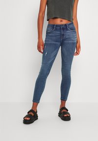 Jeans ajustados de mezclilla azul, con ligeros desgastes. Combinados con sandalias negras. Modelo con un top corto gris. Diseño sencillo.