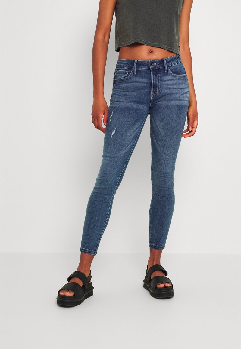 Jeans ajustados de mezclilla azul, con ligeros desgastes. Combinados con sandalias negras. Modelo con un top corto gris. Diseño sencillo.