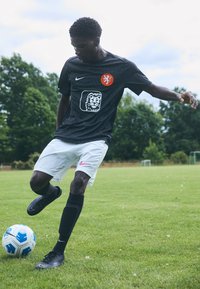 Svart fotballtrøye med oransje løveemblem, hvite shorts med rosa Nike-logo, svarte sokker og svarte fotballsko, som dribler en blå og hvit ball.