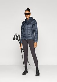 Ναυτικό μπουφάν puffer με κουκούλα, ελαφρύ και με ραμμένη υφή, συνοδευόμενο από σκουρόχρωμα κολάν και μαύρα παπούτσια πεζοπορίας, κρατώντας παγούρια πεζοπορίας.