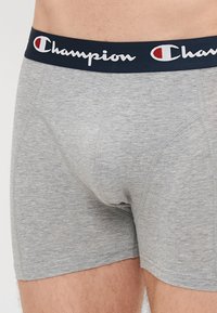 Boxer-briefs en coton gris avec une ceinture élastique marine portant le logo "Champion" en blanc. Texture lisse, longueur mi-cuisse.